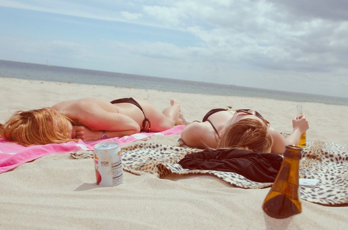 Dues noies prenent el sol en una platja, amb ampolles d'alcohol