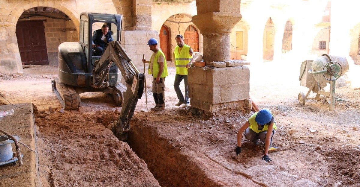 Obrers obrint una rasa amb l'excavadora mentre una arqueòloga resseguix i excava les parets d'un mur aparegut a la plaça de l'Eslgésia d'Horta de Sant Joan.
