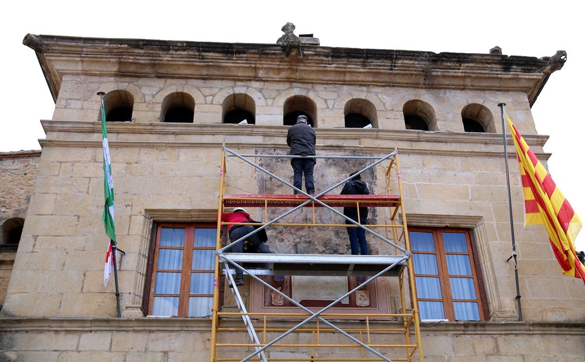 Façana de l'Ajuntament d'Horta de Sant Joan amb tres tècnics del Centre de Restauració de Bens Mobles de Catalunya prenent mostres dels mural de pintura del segle XIX.