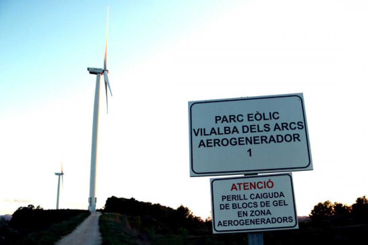 Primer pla d'un cartell senyalitzant un parc eòlic a Vilalba dels Arcs, a la Terra Alta, amb dos aerogeneradors al fons